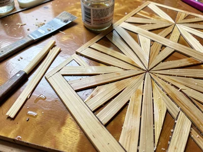 A work surface with tools — split straw strips, glue pot, and a partially inlaid
