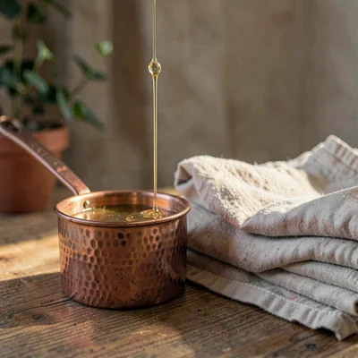 Cozy hero of a small copper butter warmer beside a stack of folded linen, a thin