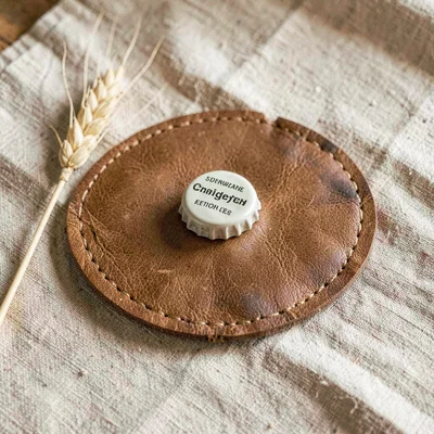 Warm flat lay of a leather coaster, an unbranded bottle cap, and a barley sprig 