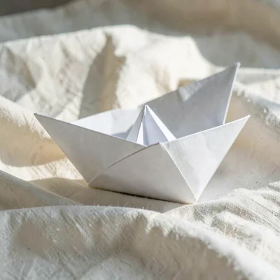 Folded paper boat resting on a bed of cream linen, sidelit, hairline crease catc