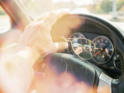 Man's weathered hand resting on a car steering wheel, dashboard reflected in avi