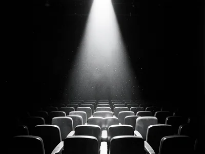 Empty theater seats receding into darkness, a single spotlight illuminating dust