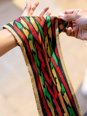 Hand lifting kente strip — fingers spread, cloth raised to light, translucent th