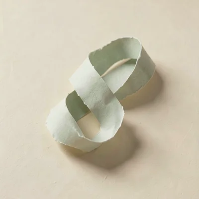 Knotted paper sculpture made from one continuous sage ribbon rests on a warm cre