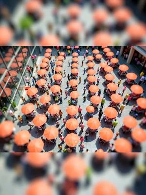 Glass-tower tilt-shift photograph downward to a busy plaza, saturated coral umbr