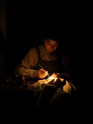 A woman sewing by lamplight, her needle catching a point of light, fabric draped