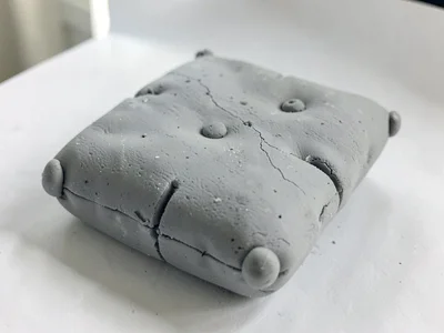Sat-upon putty-grey cushion with thumb dents along the seams and fingernail-pres