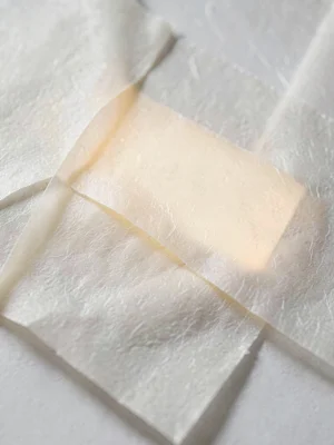 Layered-paste lift on vellum exposing a warm cream gap glowing softly between tw