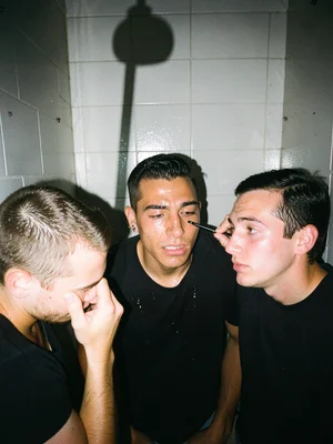 Three friends crammed into club bathroom stall, one doing another's eyeliner, fl