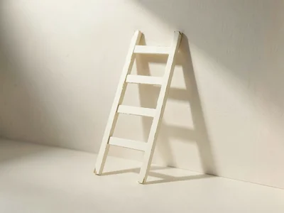 Cream paper ladder leans against an empty papercraft wall casting a long soft ti