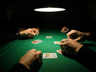 Hands dealing cards on green felt table, overhead lamp, faces in shadow