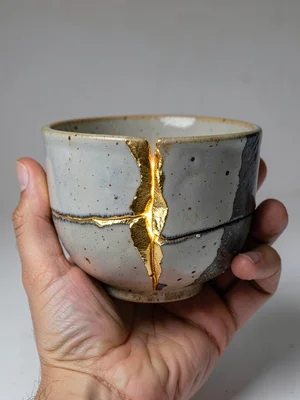 Hand cradling a repaired tea bowl — seams visible between fingers, studio light