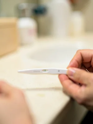 Two lines on pregnancy test in sharp focus, bathroom counter a soft blur, hands 