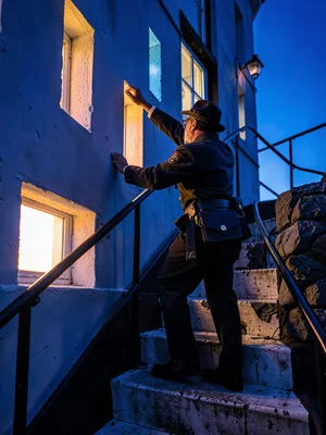 Lighthouse keeper climbing spiral stairs at blue hour, window slits alternating 