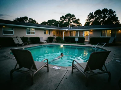 A roadside motel pool at dusk, turquoise water, two deck chairs, a single swimme