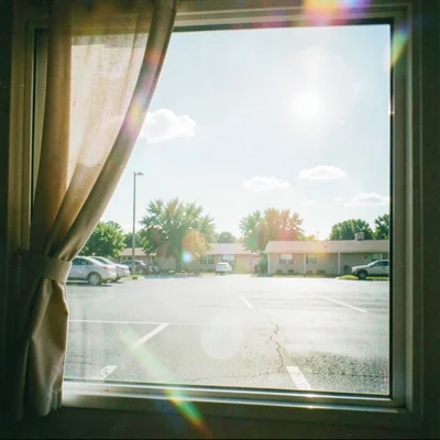 Curtain of a motel window pulled half open onto a midday parking lot with two sl