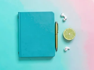 Top-down knolled arrangement of a teal notebook, brass pen, AirPod-style buds, a