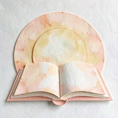 Layered papercraft of a sun rising behind a stylized open book, warm peach and o