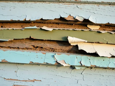 Peeling paint on clapboard siding revealing five layers: current pale blue, 1970