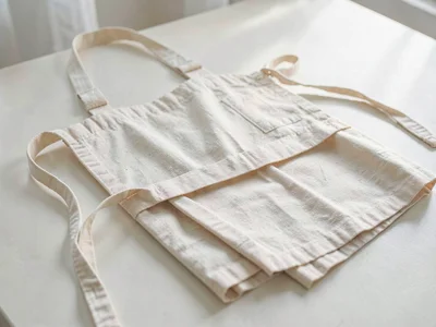 The folded raw cotton apron lies flat showing soft canvas weave and long woven w