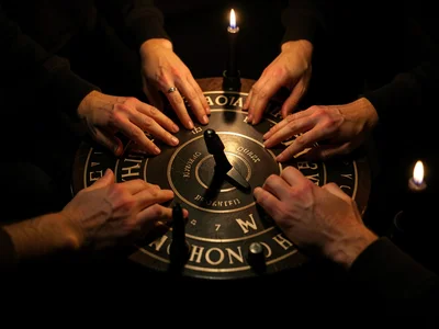 Four pairs of hands around a ouija board lit by black candles, the planchette mi