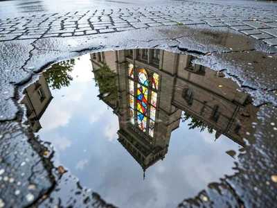 Stained glass window reflected in church courtyard puddle, fragmented color on g
