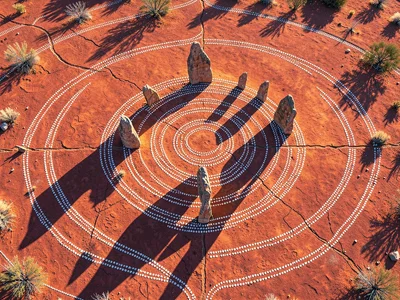 Standing stones at desert dawn from above — long shadows radiating outward acros