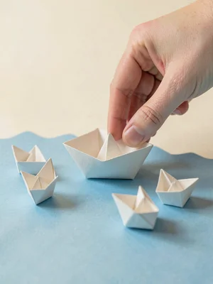 A papercraft hand reaching down to lift a single folded paper boat from a flat b