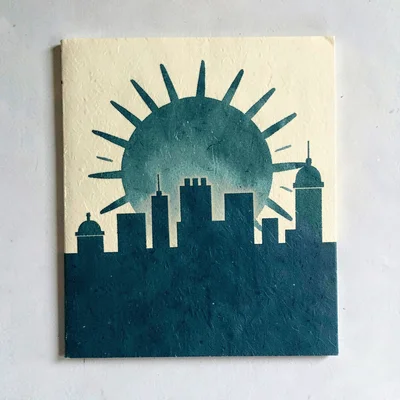 Two-color riso cover of a stamper-shaped sun rising over a paper city, deep teal