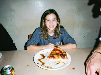 Half-eaten slice of pizza on a paper plate at a folding table, grease pooling, s