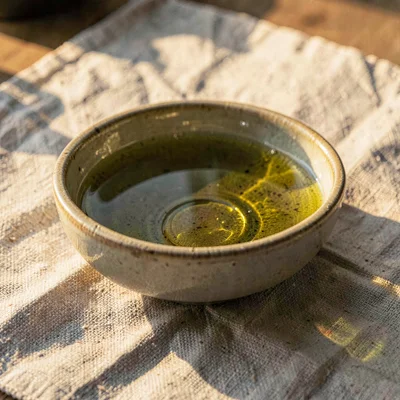 A small ceramic dipping bowl of golden-green oil on raw linen, warm afternoon gl