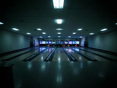 An empty bowling alley at closing time, all sixteen lanes dark except the approa