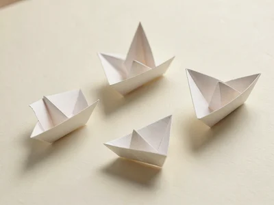 Three identical paper boats float on a flat cream surface with one boat tilted s