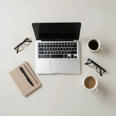 Overhead flat lay of a work-from-home setup: laptop, notebook, pens, glasses, co