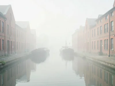 Slack canal between brick warehouses, one barge half-vanished in vapor, water re