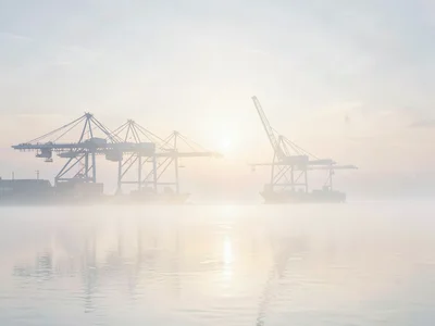 Quayside loading cranes silhouetted in dawn vapor, the river reduced to a flat p
