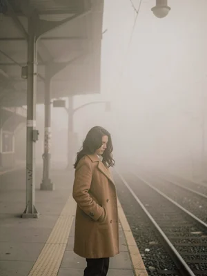 Woman in camel coat standing at a train platform, fog erasing everything beyond 