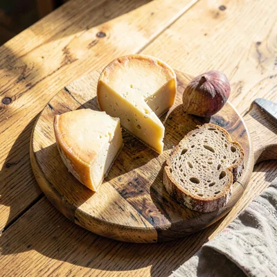 Warm overhead of a cheese board with three wedges, fig and bread on a sunlit oak