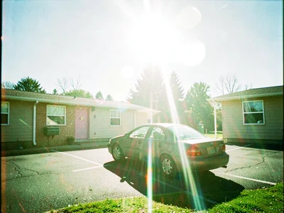 Small motel parking lot at noon with a single parked sedan and a long lens flare