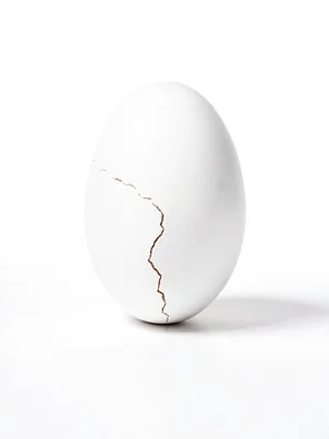 Single white egg balanced on end on white surface, hairline crack just visible, 