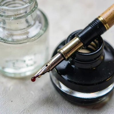 Macro of a dip pen resting against a glass ink well, ink beading at the nib