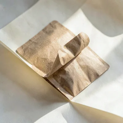 Single folded kraft pouch unfurling against a cream paper sweep, soft directiona