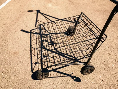 The shadow of a shopping cart cast onto parking lot asphalt at noon, the wire me
