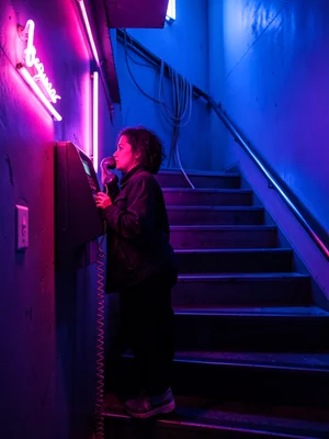Person on payphone in neon-lit stairwell, cable coiling, emotional conversation