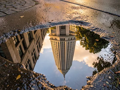 Building facade reflected in sidewalk puddle, inverted skyscraper reaching down 