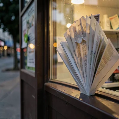 Paper rosette pinned to a kiosk wall echoing a folded fan of pages, dusk-warm ke