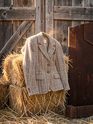 Chanel tweed jacket draped on hay bale, weathered barn wood behind, glamour meet