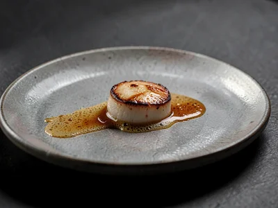 Hero plate of a single seared scallop trailing brown-butter foam on smoke-grey c