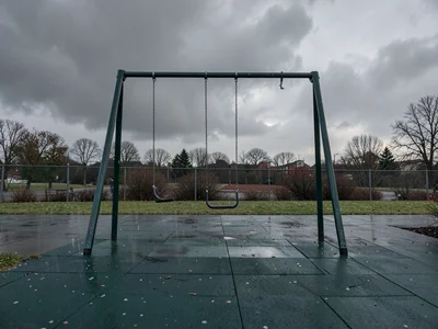 An empty playground under heavy grey sky, a single swing still moving from its l