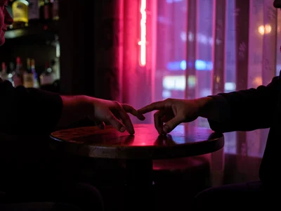 Hands reaching across small table in dim bar, neon light through curtain, intima